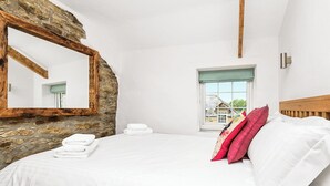 A bedroom with a stone wall, a wooden headboard, a bed with white linens, a mirror, and a window with a view of a house.