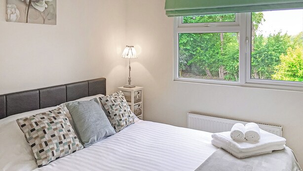 A neatly made bed with pillows, a bedside lamp, a radiator, and a window with a view of greenery.