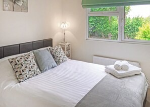 A neatly made bed with pillows, a bedside lamp, a radiator, and a window with a view of greenery.
