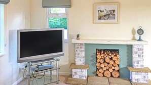 A living room with a flat-screen TV on a glass stand, a fireplace with stacked logs, and a framed picture above it.