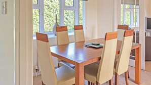 A dining area with a wooden table and six chairs, a vase on the windowsill, and a television in the background.