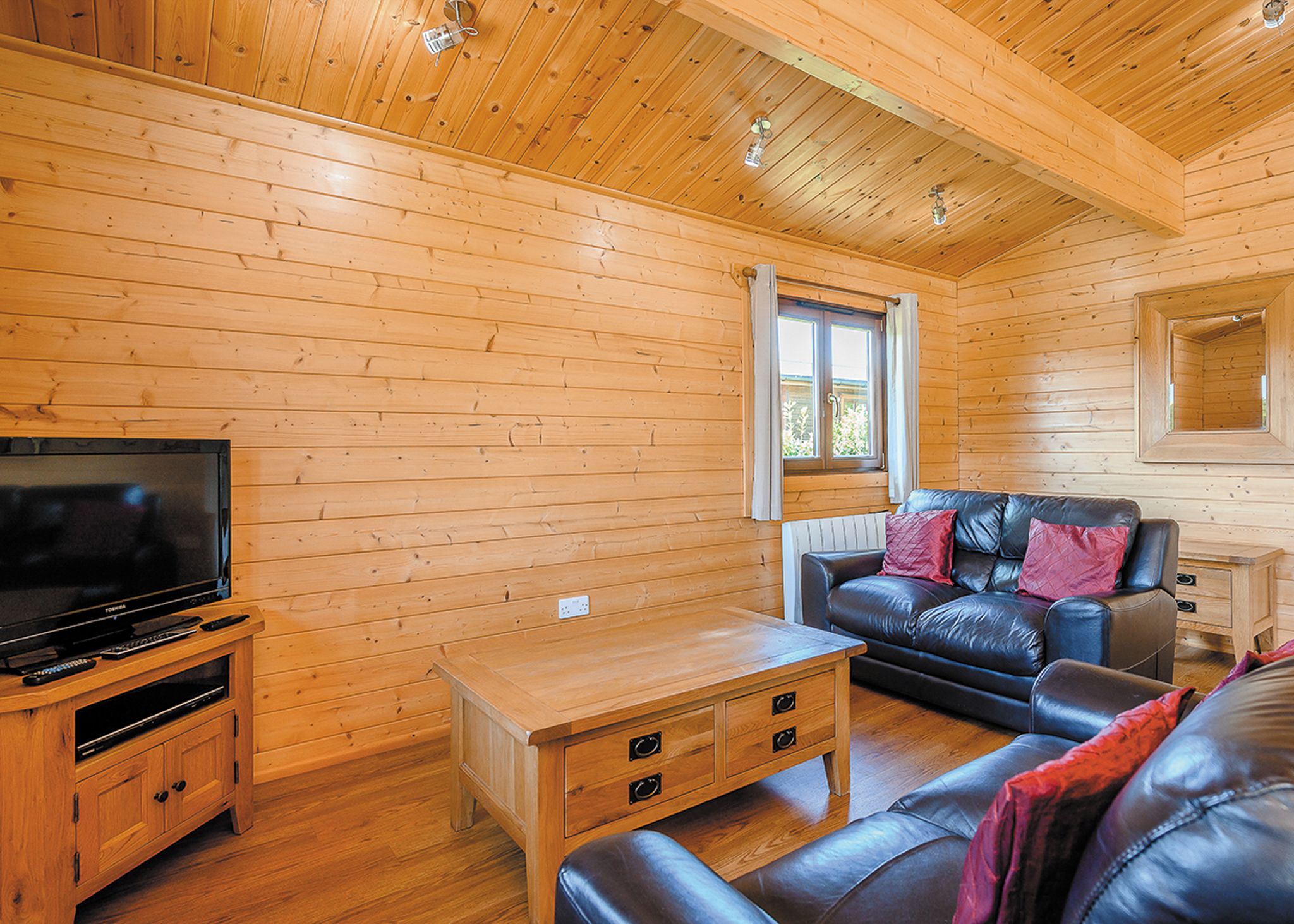 A wooden cabin interior with a television on a wooden stand, a leather sofa, a wooden coffee table, and a window with curtains.