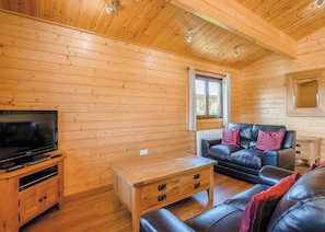 Ein Interieur einer Holzhütte mit einem Fernseher auf einem Holzständer, einem Ledersofa, einem Holztisch und einem Fenster mit Vorhängen.