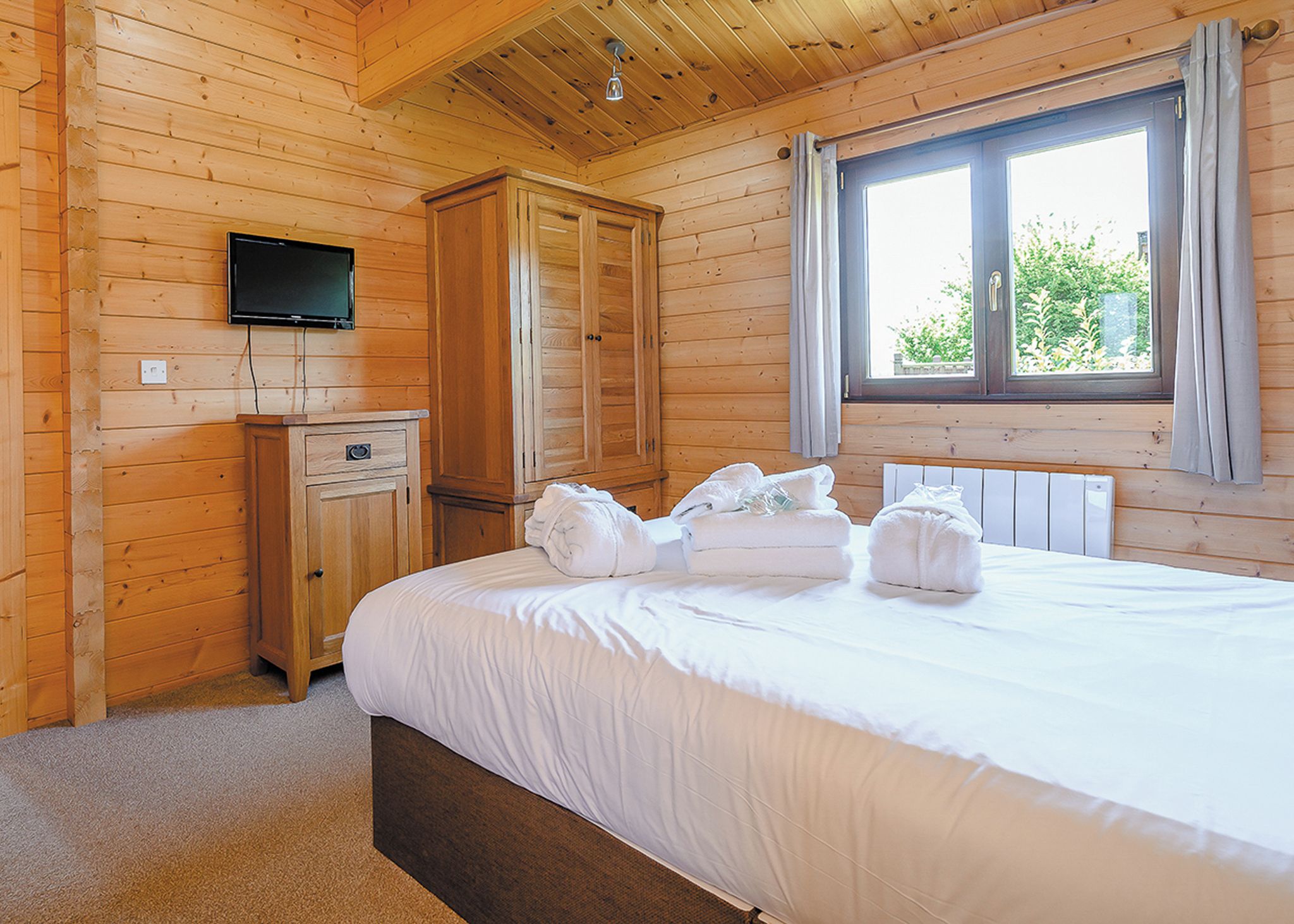 A wooden-paneled room with a bed, a television mounted on the wall, and a window with curtains.