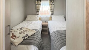 Una habitación estrecha con dos camas, una ventana con cortinas estampadas y un enchufe visible.