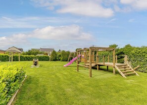 A wooden playground with a slide and climbing structures in a grassy yard with hedges and a house in the background. - Luxurious and stylish modern lodge with one master double and one twin. (Haswell, Nr Durham)