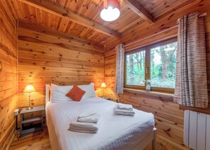 Camera da letto in una baita di legno con un letto, due lampade sui comodini, una finestra con tende e vista sugli alberi.