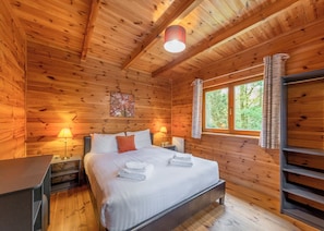 A wooden cabin bedroom with a bed, two bedside lamps, a window with curtains, and a painting on the wall.
