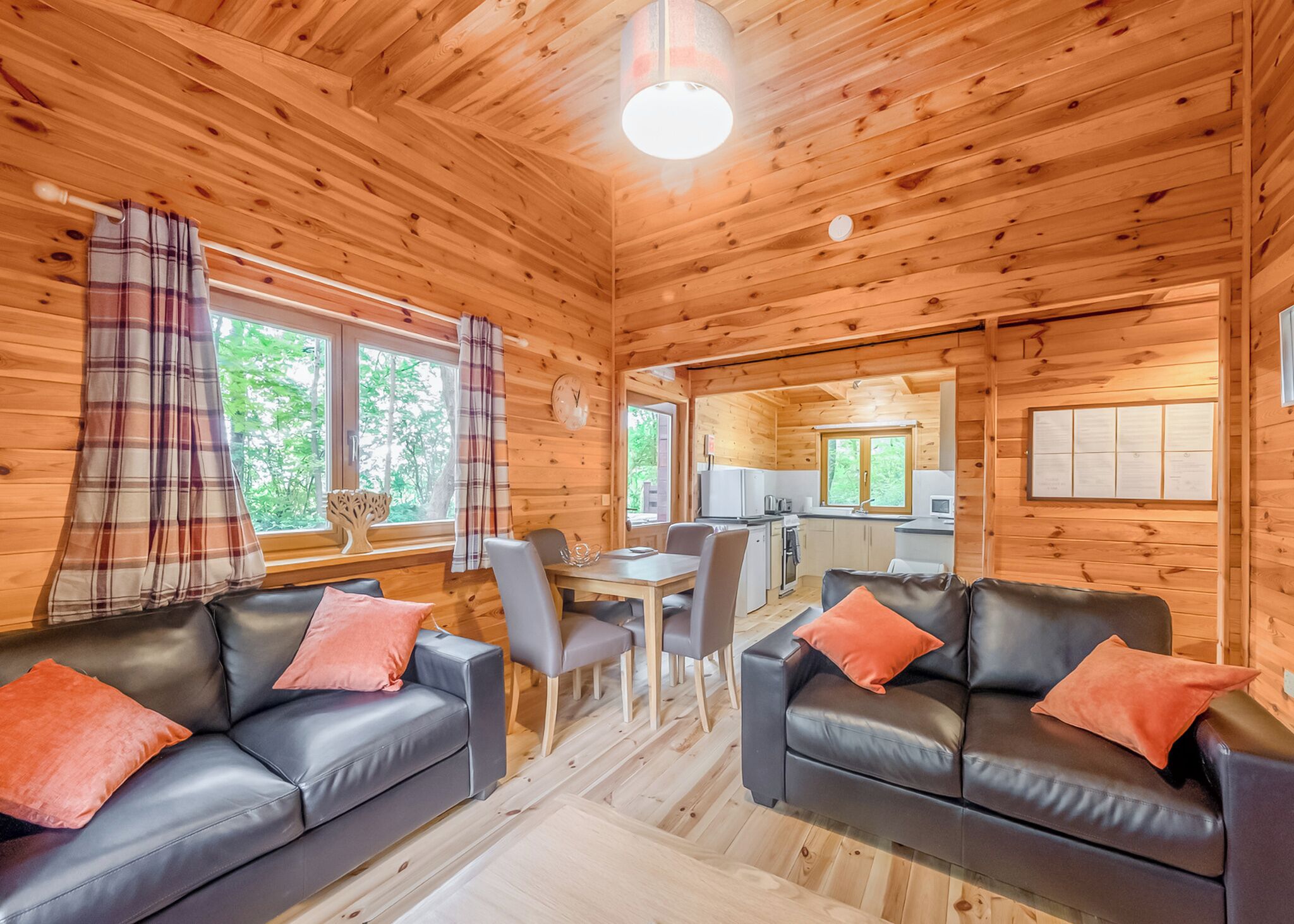 A cabin with wooden walls, a dining area, two black leather sofas with orange cushions, and a kitchen in the background.