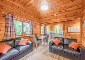 A cabin with wooden walls, a dining area, two black leather sofas with orange cushions, and a kitchen in the background.