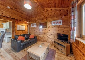 Interior de una cabaña de madera con sofá, televisión, armarios de madera y una zona de cocina.