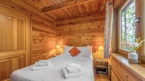 A wooden cabin room with a bed, two bedside lamps, a window with curtains, and a vase with flowers. - Woodland Willow Lodge (Ellesmere)