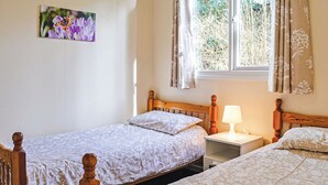 A bedroom with two wooden beds, a nightstand with a lamp, a window with curtains, and a painting on the wall. - 2 bedroom accommodation in Ruthernbridge, Nr Bodmin (Ruthernbridge, Nr Bodmin)