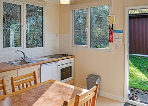 Une cuisine équipée d’un évier, d’une cuisinière et d’un four, une table à manger avec des chaises, et de grandes fenêtres donnant sur un paysage verdoyant.