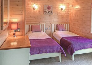 A wooden cabin-style room with two beds, a nightstand, a lamp, and a wall-mounted light fixture.