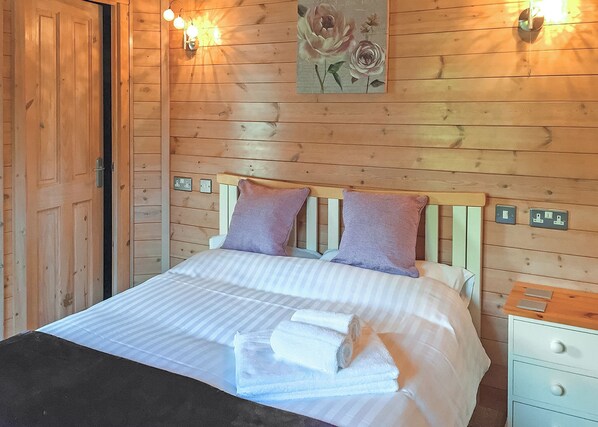 A wooden-paneled room with a bed, two pillows, a painting, and wall-mounted lights.