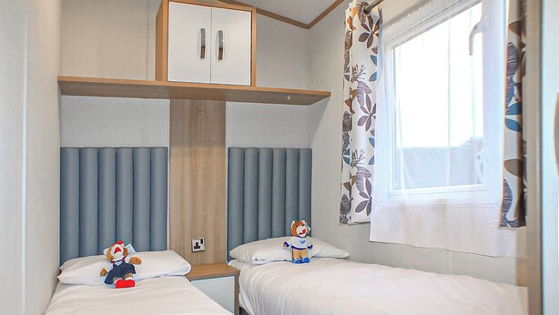 A bedroom with two beds, a wardrobe, a window with curtains, and a teddy bear on one bed. - 12ft-wide caravan with one double and one twin. (Silloth)