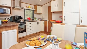 A compact kitchen with a microwave, stove, and various appliances. There is a dining table with breakfast items. - 12ft-wide caravan with one double and one twin. (Silloth)