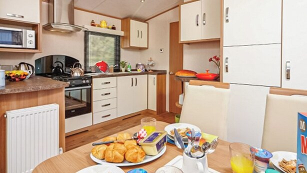 A compact kitchen with a microwave, stove, and various appliances. There is a dining table with breakfast items. - 12ft-wide caravan with one double and one twin. (Silloth)