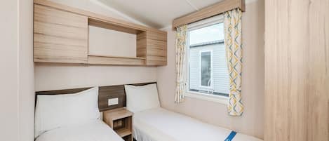 Une pièce de taille modeste, comprenant deux lits, une petite table de chevet, une fenêtre avec des rideaux et des étagères en bois intégrées.