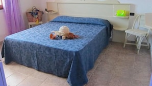 A bedroom with a bed covered in a blue bedspread, a wicker chair, a small shelf with a green object, and a basket on a chair. - LE CASTELLA RESIDENCE APP. 7 (Santa Domenica)
