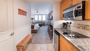 Une cuisine compacte équipée d’un four à micro-ondes, d’une cuisinière et d’un évier. Un banc en bois se trouve près de la porte, et on aperçoit un lit au fond de la pièce.