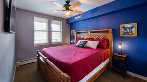 Une chambre à coucher avec un lit en bois, un ventilateur de plafond, deux fenêtres équipées de stores et une table de chevet avec une lampe.