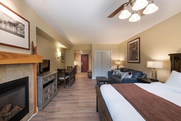A hotel room with a fireplace, a bed, a sofa, a desk with chairs, and a ceiling fan.
