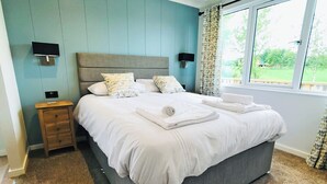 Ein ordentlich bezogenes Bett mit Kissen und Handtüchern, ein Nachttisch mit Fernbedienung und ein Fenster mit Vorhängen.