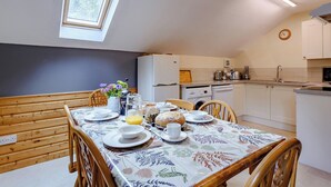 A dining area with a table set for four, a window, a ceiling light, and a kitchen in the background. - 3 bedroom accommodation in Webbery, Bideford (Webbery, Bideford)