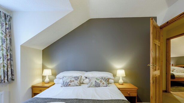 A bedroom with a sloped ceiling, a bed with white linens and floral pillows, two bedside lamps, and a wooden door leading to another room. - 3 bedroom accommodation in Webbery, Bideford (Webbery, Bideford)
