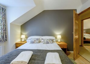A bedroom with a sloped ceiling, a bed with white linens and floral pillows, two bedside lamps, and a wooden door leading to another room. - 3 bedroom accommodation in Webbery, Bideford (Webbery, Bideford)
