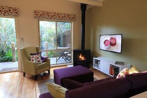 Een gezellige woonkamer met een bank, fauteuil en poef, een open haard, een televisie en een groot raam met uitzicht op groen.