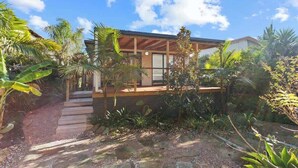 A modern house with a wooden deck, surrounded by tropical plants and a stone pathway.