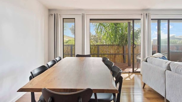 A modern dining room with a long wooden table and black chairs, a grey sofa, and a sliding glass door leading to a balcony with a view of greenery.