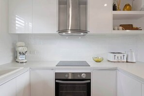 Cocina compacta con armarios blancos, horno integrado, campana extractora de acero inoxidable, cafetera y tostadora.