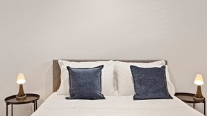 Una cama con ropa de cama blanca y dos almohadas azules, flanqueada por dos mesitas de noche con lámparas.