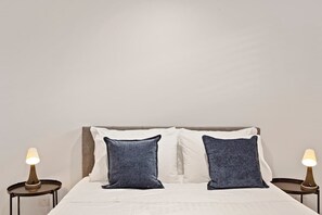 Una cama con ropa de cama blanca y dos almohadas azules, flanqueada por dos mesitas de noche con lámparas.
