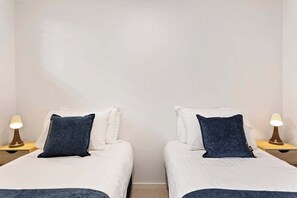 A hotel room with two beds, each with a white mattress and a dark blue pillow. There are bedside lamps on wooden nightstands.