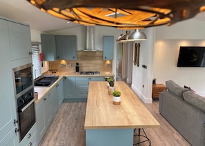 A modern kitchen with a central island, built-in oven, and a dining area with a grey sofa. - Experience the ultimate retreat in this stunning woodland lodge. (Chipping)