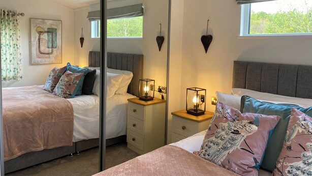 A bedroom with a mirrored closet, a bed with multiple pillows, bedside tables with lamps, and a window with a view of trees. - Experience the ultimate retreat in this stunning woodland lodge. (Chipping)