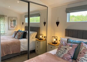 A bedroom with a mirrored closet, a bed with multiple pillows, bedside tables with lamps, and a window with a view of trees.