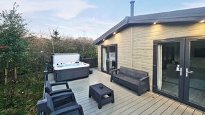 A hot tub on a wooden deck with outdoor furniture, a glass door, and a chimney. - Experience the ultimate retreat in this stunning woodland lodge. (Chipping)