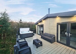 A hot tub on a wooden deck with outdoor furniture, a glass door, and a chimney. - Experience the ultimate retreat in this stunning woodland lodge. (Chipping)