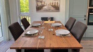 A dining table set for four with chairs, a painting above, and a view of greenery outside. - Experience the ultimate retreat in this stunning woodland lodge. (Chipping)