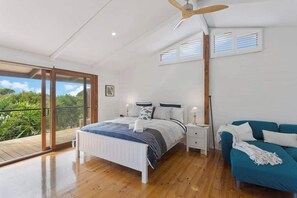 Un dormitorio con cama, mesitas de noche, ventilador de techo, ventana con persianas y balcón con vistas a la vegetación.