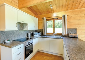 A compact kitchen with white cabinets, a built-in oven, a microwave, a toaster, and a window with curtains. - Luxurious solid log lodge. (Wenhaston, Halesworth)