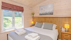 A bedroom with a bed, two bedside tables, a window with a view, and a painting on the wall. - Luxurious lodge with stunning countryside views. (Ashover)