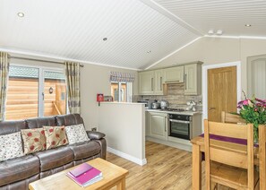A compact living space with a sofa, a coffee table, a kitchen area with cabinets and appliances, and a wooden door. - Woodland Retreat Plus (Water Yeat, Nr Coniston)