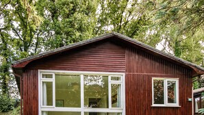 A wooden cabin with large windows, a porch with a green sofa and a small table, surrounded by lush greenery.